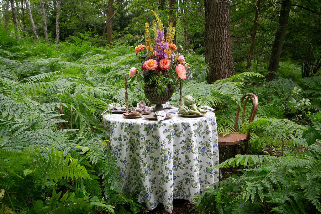 A round table set in a wood dressed with a full lenght cotton block printed tablecloth and a large urn of flowers 