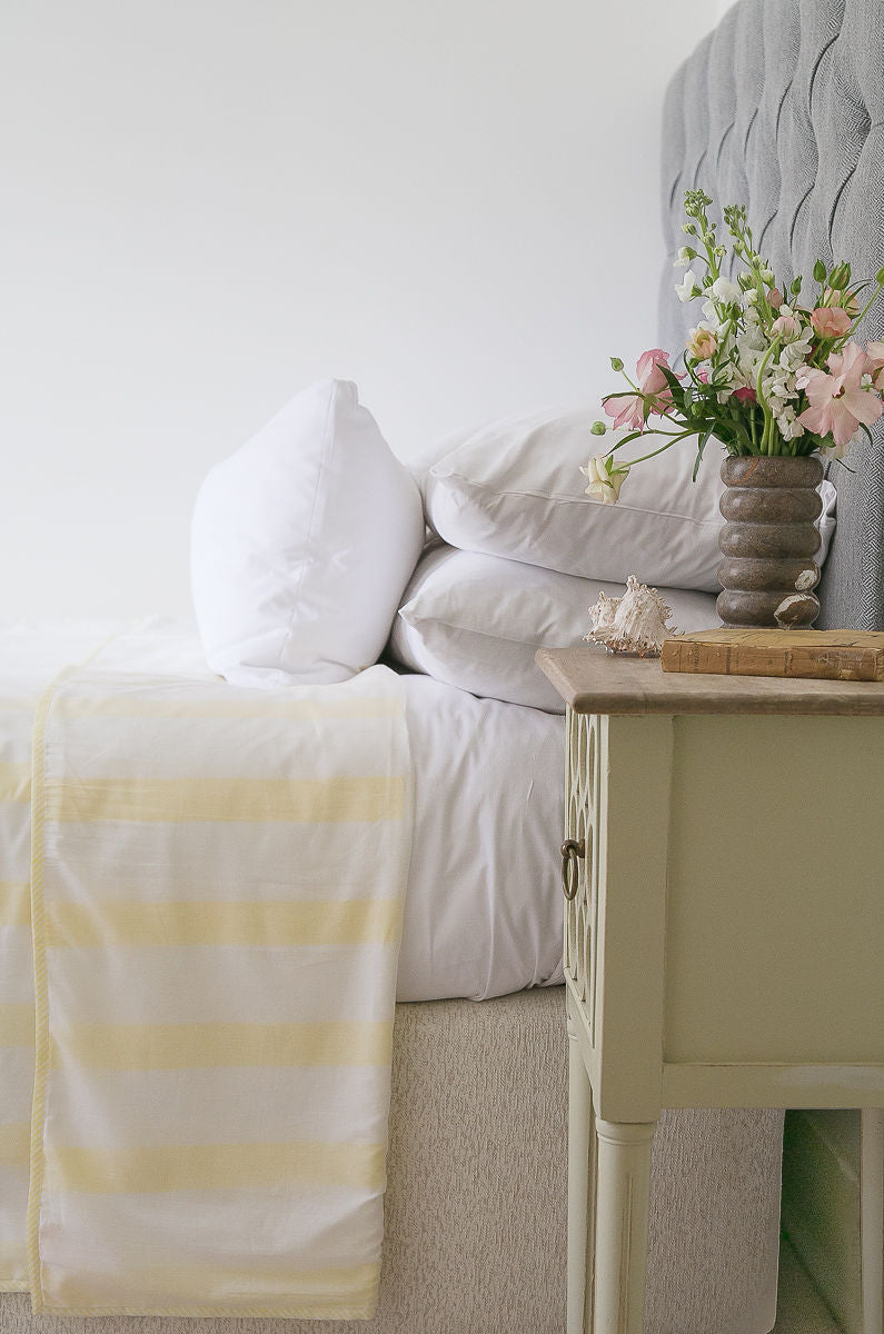 Bedroom with a bed featuring a yellow and white striped Dohar blanket, pillows, and a nightstand with flowers.