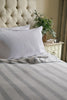 Neatly made bed with striped Sleep Dohar and pillows, nightstand with books and plant in the background.