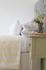 Bedroom with a bed featuring a yellow and white striped Dohar blanket, pillows, and a nightstand with flowers.