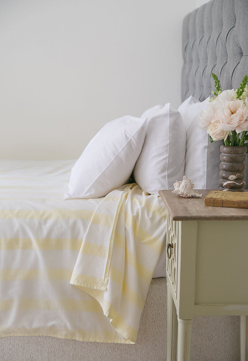 Bed with white bedding and yellow striped Dohar blanket next to a nightstand with flowers.