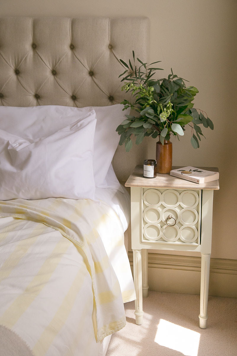 a bed with a striped Sleep Dohar and a bedside cabinet with a vase of foliage and a book.