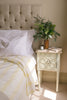 a bed with a striped Sleep Dohar and a bedside cabinet with a vase of foliage and a book.