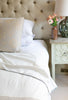 Neatly made bed with white bedding  including a Dohar and decorative pillows, nightstand with lamp and flowers.