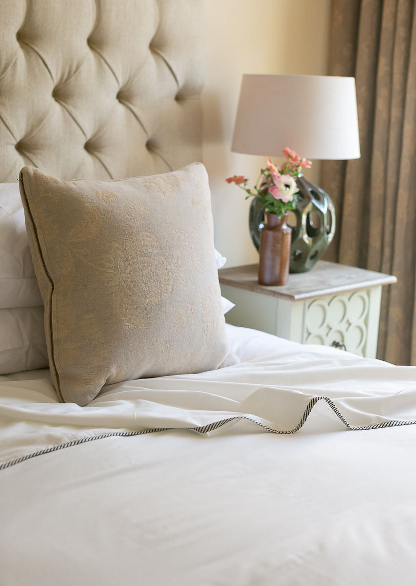 Neatly made bed with a cotton Dohar and a tufted headboard, lamp, and vase on a nightstand.