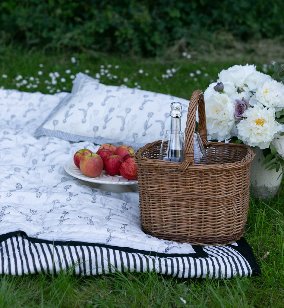 block printed quilt used as a picnic blanket with a plate of Apples a vase of Peonies and a cane picnic basket.